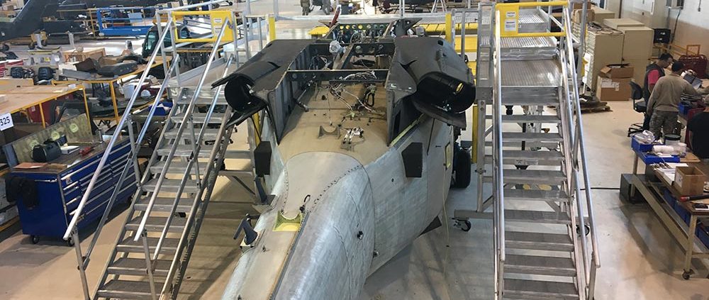 A military jet undergoing maintenance in a hangar with technicians working around it.