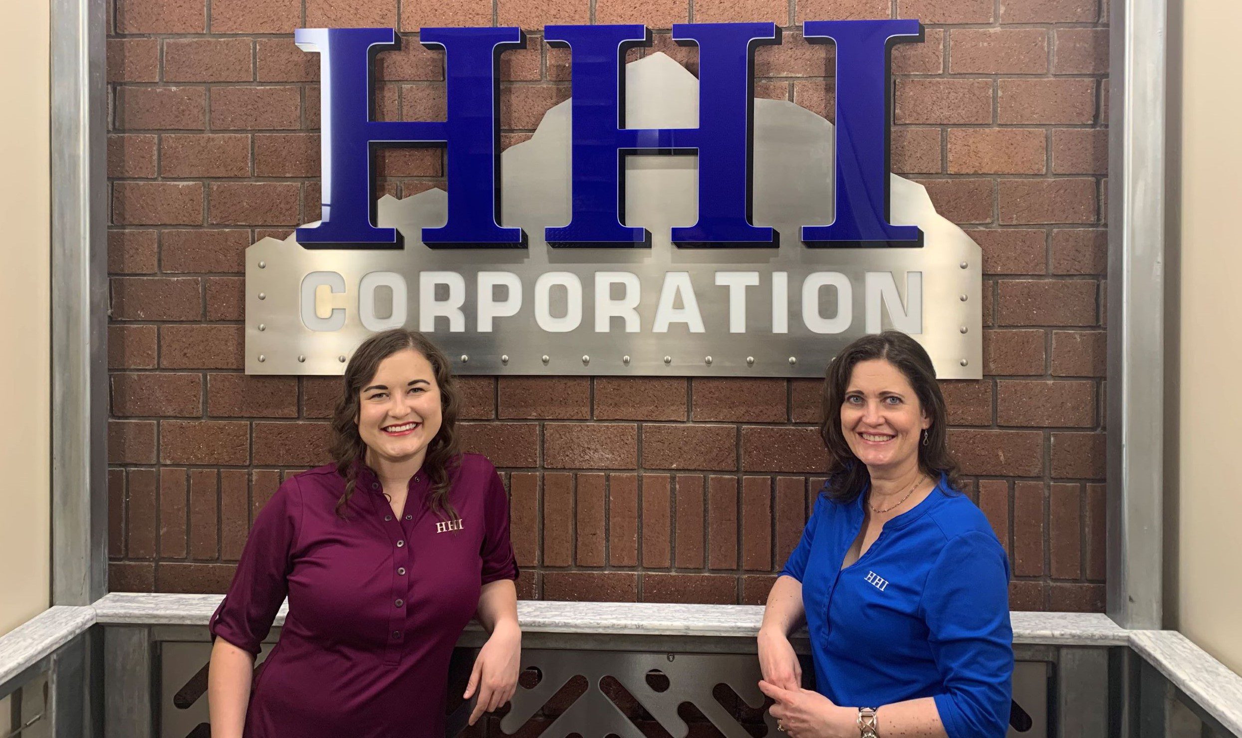 Women posing by HHI Corporation sign.